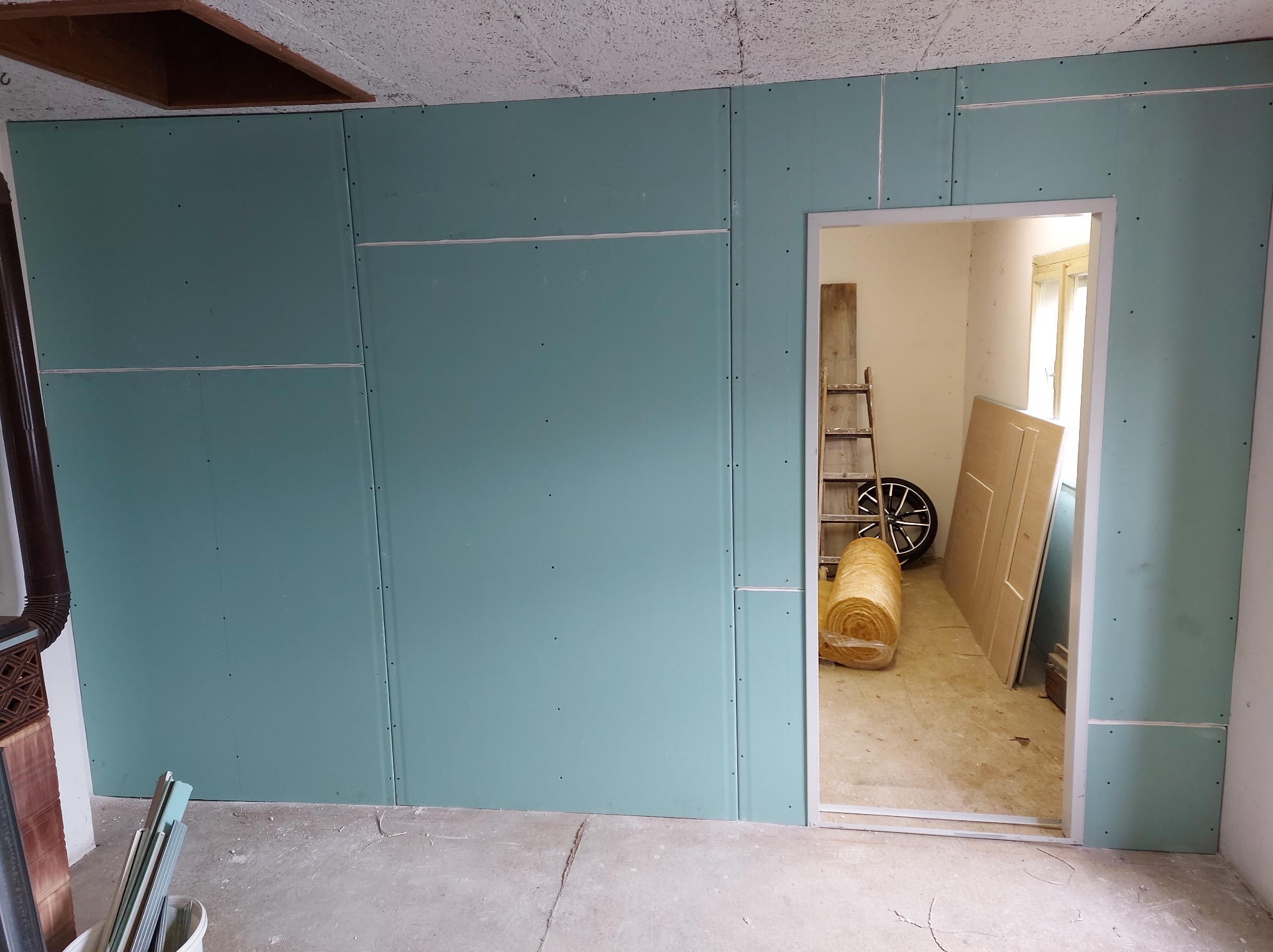 Montaż płyt gipsowo-kartonowych na ścianie, widoczne łączenia płyt i otwór drzwiowy z widokiem na drabinę i izolację w rolce.