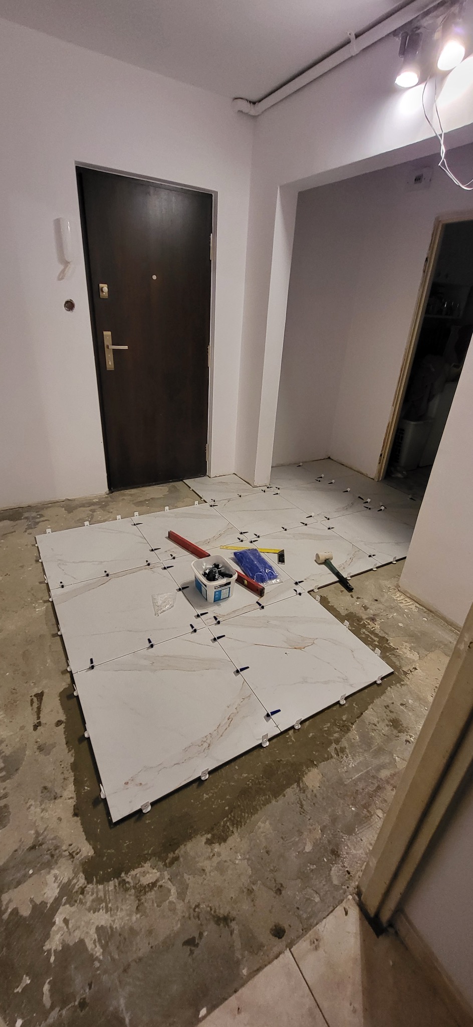 Układanie jasnych, marmurkowych płytek podłogowych w korytarzu, widoczne narzędzia glazurnicze i poziomica, fuga jeszcze nie położona.