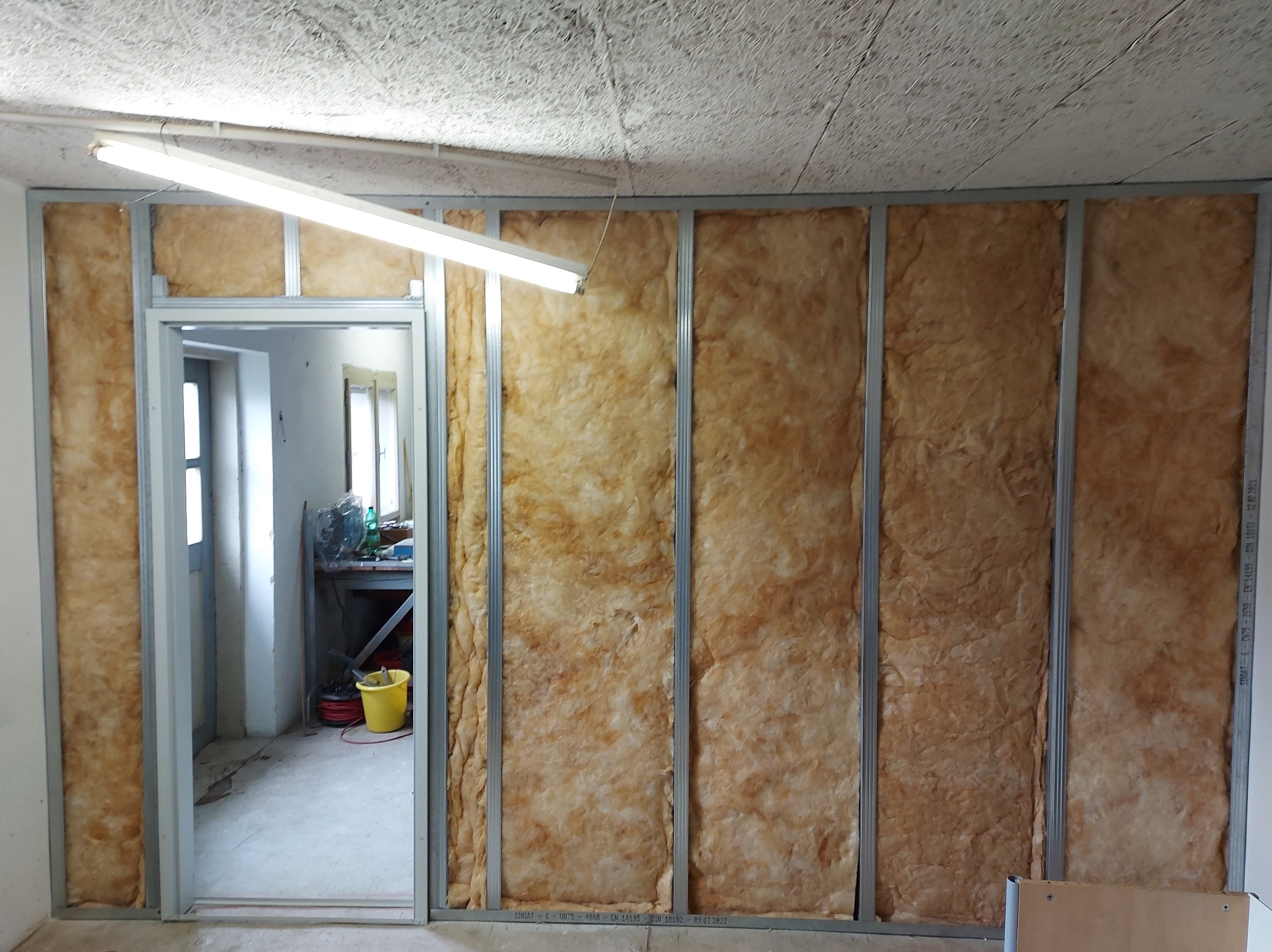 Ściana w trakcie izolacji wełną mineralną, widoczne profile metalowe i fragment otwartych drzwi, w tle żółte wiadro