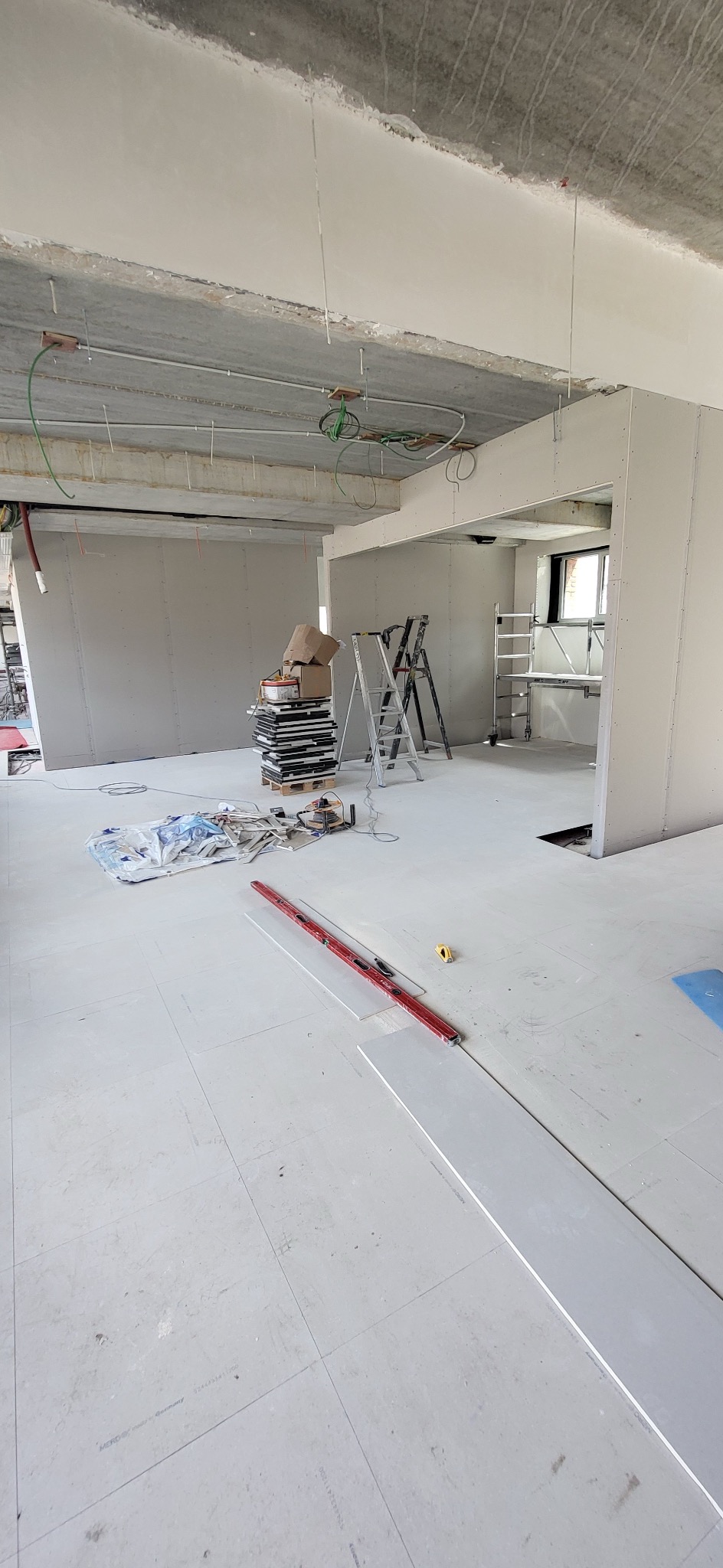 Wnętrze w trakcie remontu: widoczne płyty gipsowo-kartonowe na ścianach, surowy betonowy sufit z instalacjami, ułożone panele podłogowe, drabiny i narzędzia.