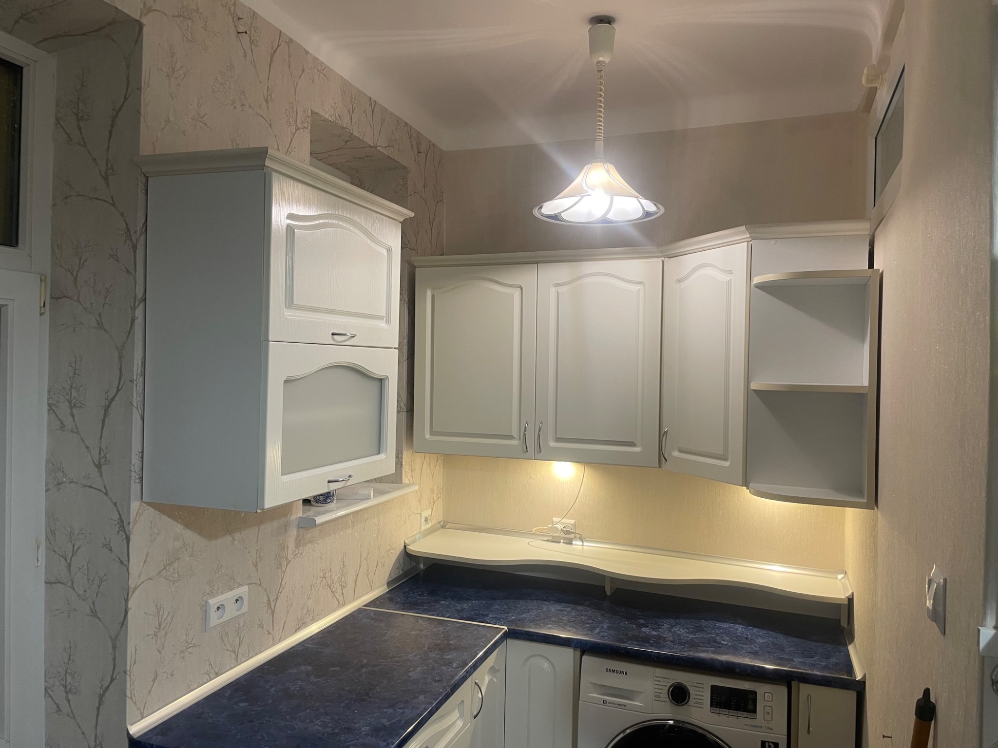 Jasna kuchnia z białymi szafkami, niebieskim blatem i wzorzystą tapetą, oświetlona wiszącą lampą i podświetleniem pod szafkami.