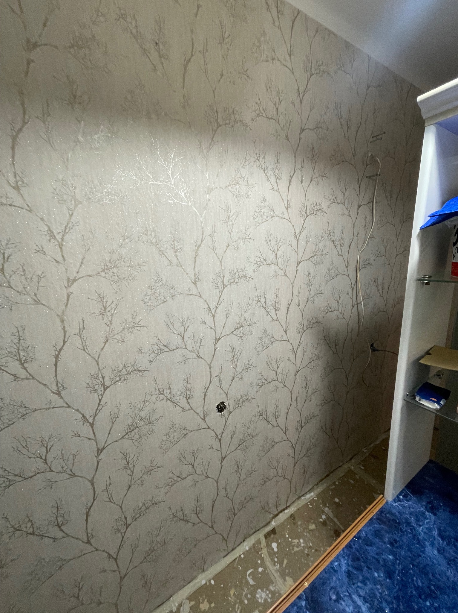 Beżowa tapeta z motywem drzew w pokoju podczas remontu, widoczne niedoskonałości przy podłodze i wystający kabel ze ściany.