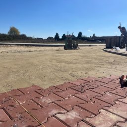 PoliTech - Czerwona kostka brukowa typu 'I', częściowo ułożona na piaskowym podłożu, z widoczną linią wyznaczającą kierunek, w tle wózek i pracownik przygotowujący kolejne elementy.