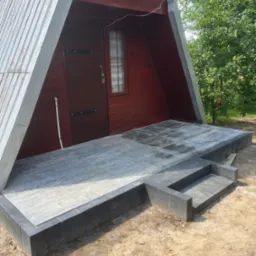 Nowoczesny taras z szarej kostki brukowej przed domkiem w stylu A-frame. Schody wejściowe wykończone tą samą kostką. Widok na brązowe drzwi wejściowe.