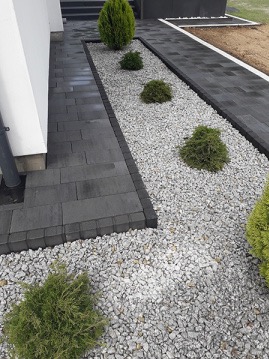 Nowoczesna ścieżka z ciemnoszarej kostki brukowej prowadząca do domu, obramowana kostką krawężnikową, z pasem wysypanym białym grysem i obsadzonym kulistymi krzewami iglastymi.