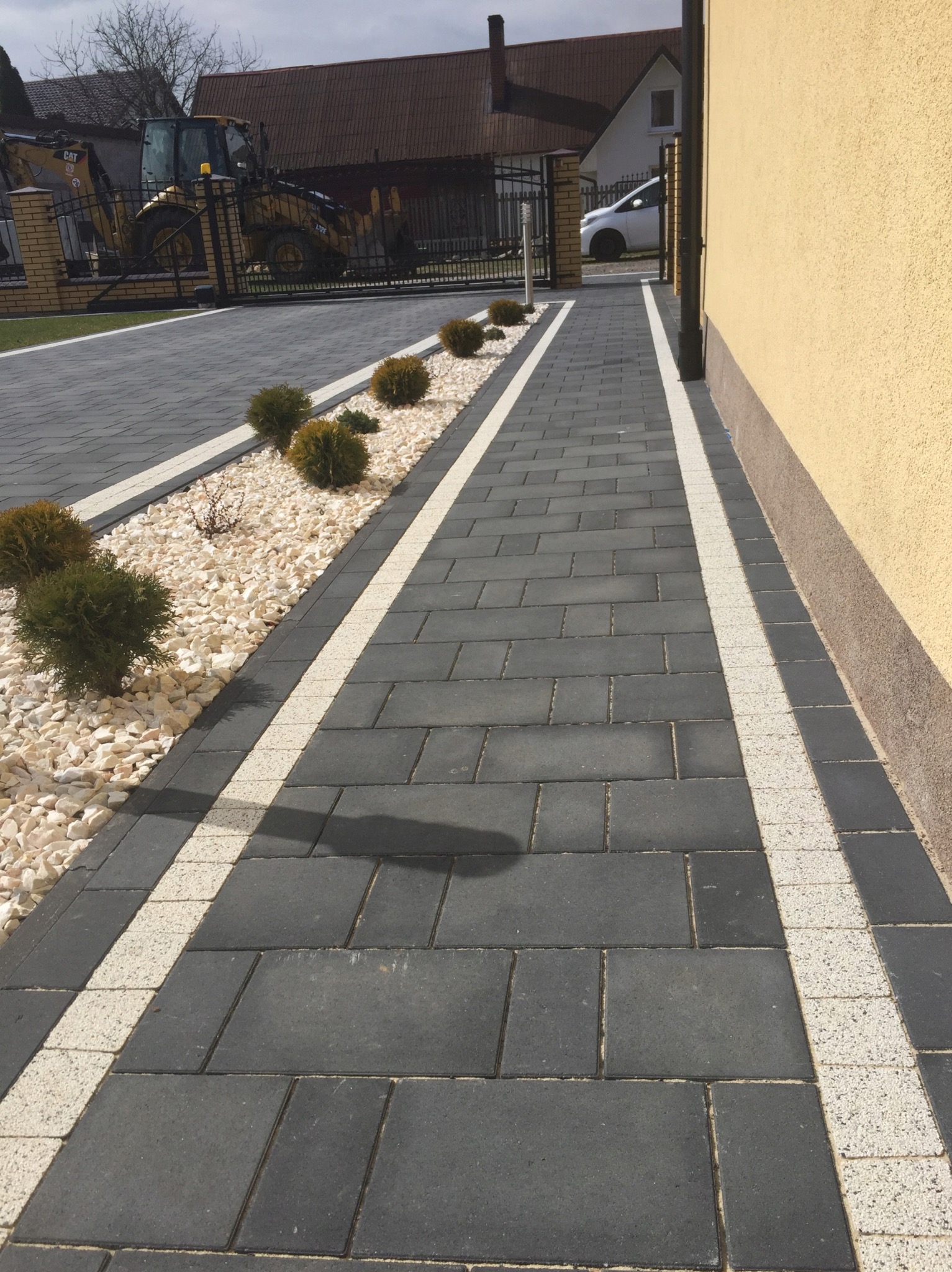Nowoczesny podjazd z ciemnoszarej kostki brukowej, obramowany jasnymi pasami i białymi kamieniami, z dekoracyjnymi krzewami i widocznym w tle traktorem budowlanym.