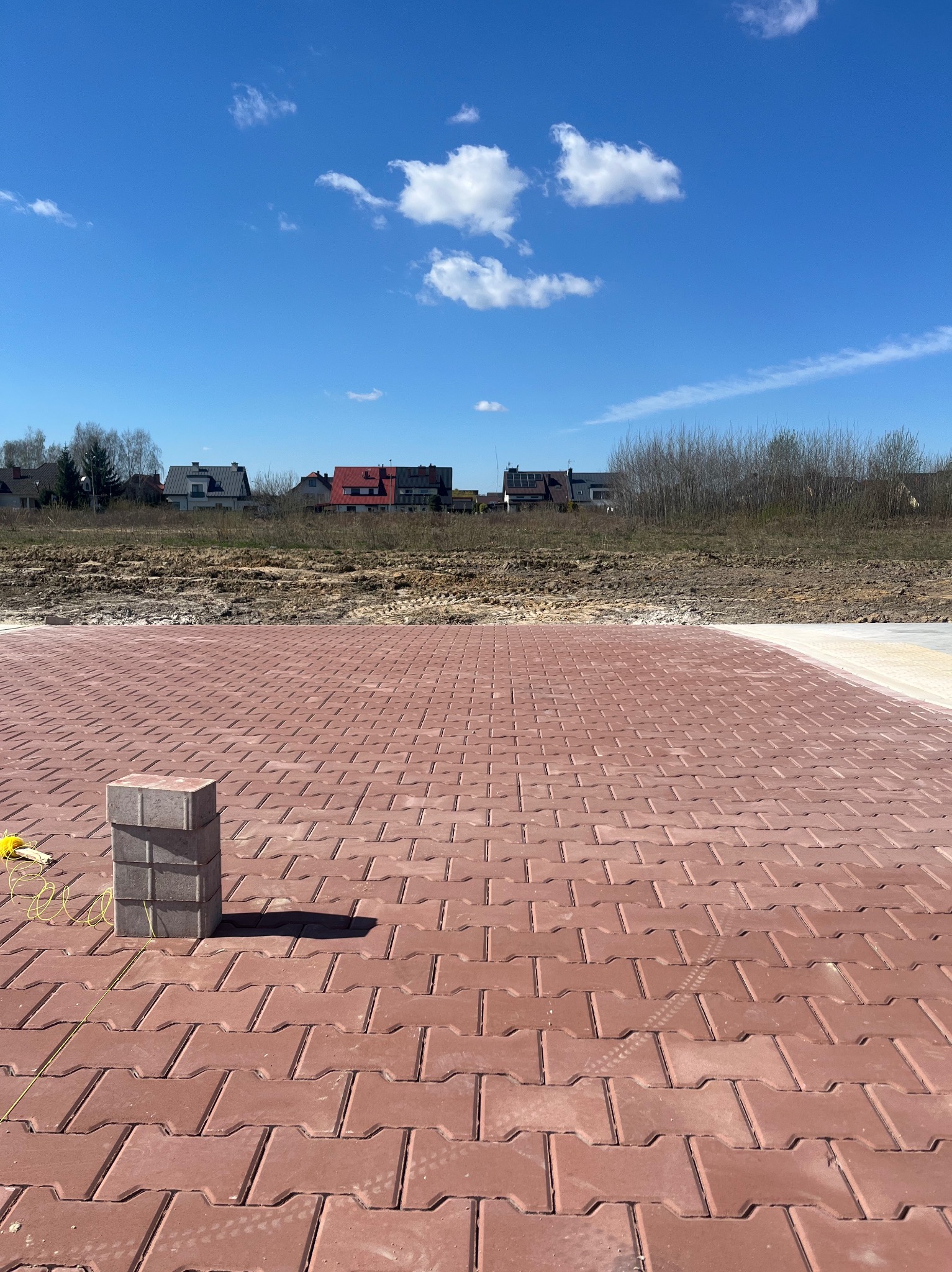 Czerwona kostka brukowa w trakcie układania, stos kostki na ułożonej powierzchni, w tle domy i błękitne niebo z chmurami.