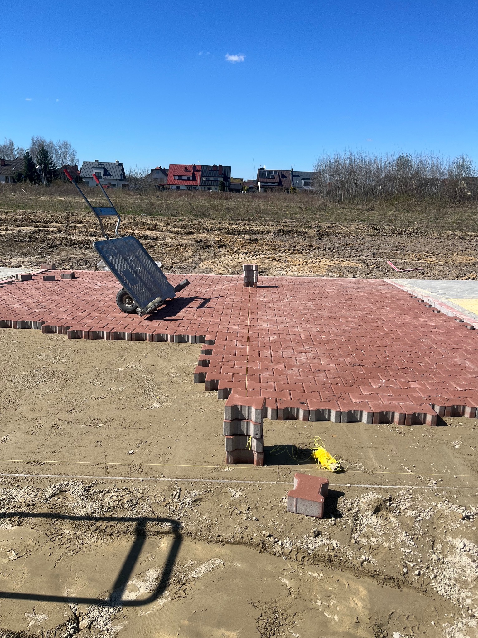 Częściowo ułożona czerwona kostka brukowa na piaszczystym podłożu, wózek transportowy z kostką w tle, widoczne żółte sznurki wyznaczające linię, w tle zabudowa jednorodzinna pod błękitnym niebem.