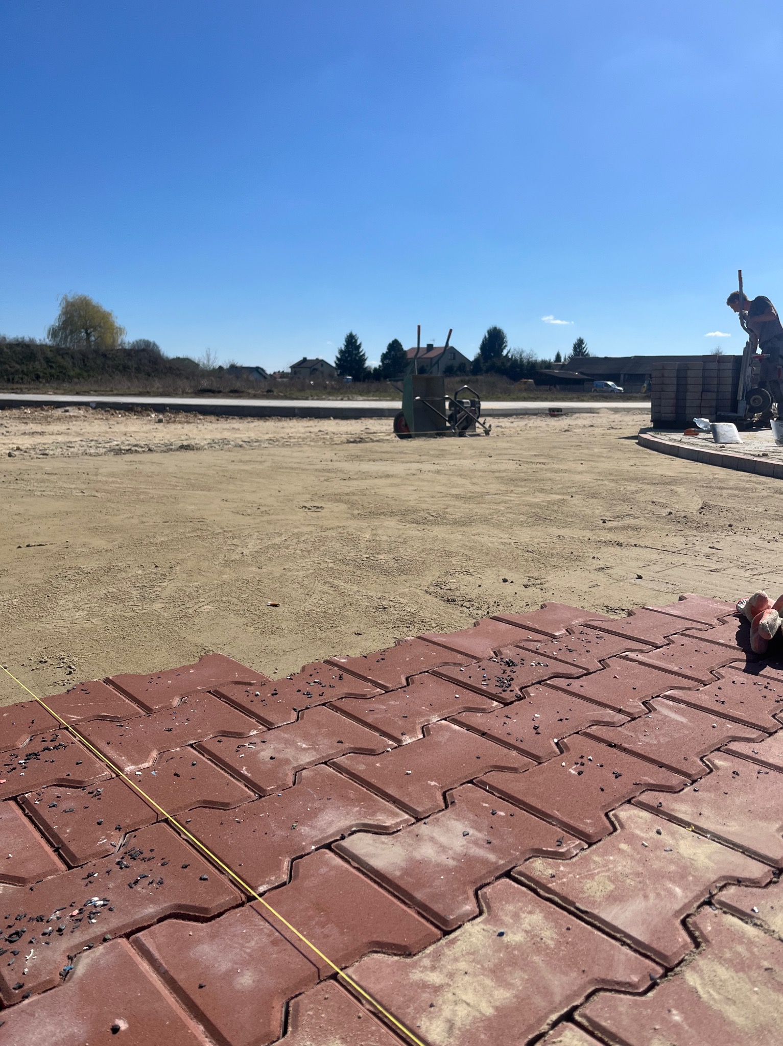 Czerwona kostka brukowa typu 'I', częściowo ułożona na piaskowym podłożu, z widoczną linią wyznaczającą kierunek, w tle wózek i pracownik przygotowujący kolejne elementy.