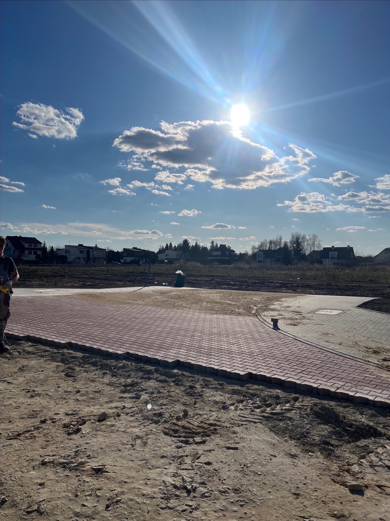 Częściowo ułożona, czerwona kostka brukowa na posesji z widocznym pracownikiem i błękitnym niebem z chmurami w słoneczny dzień.