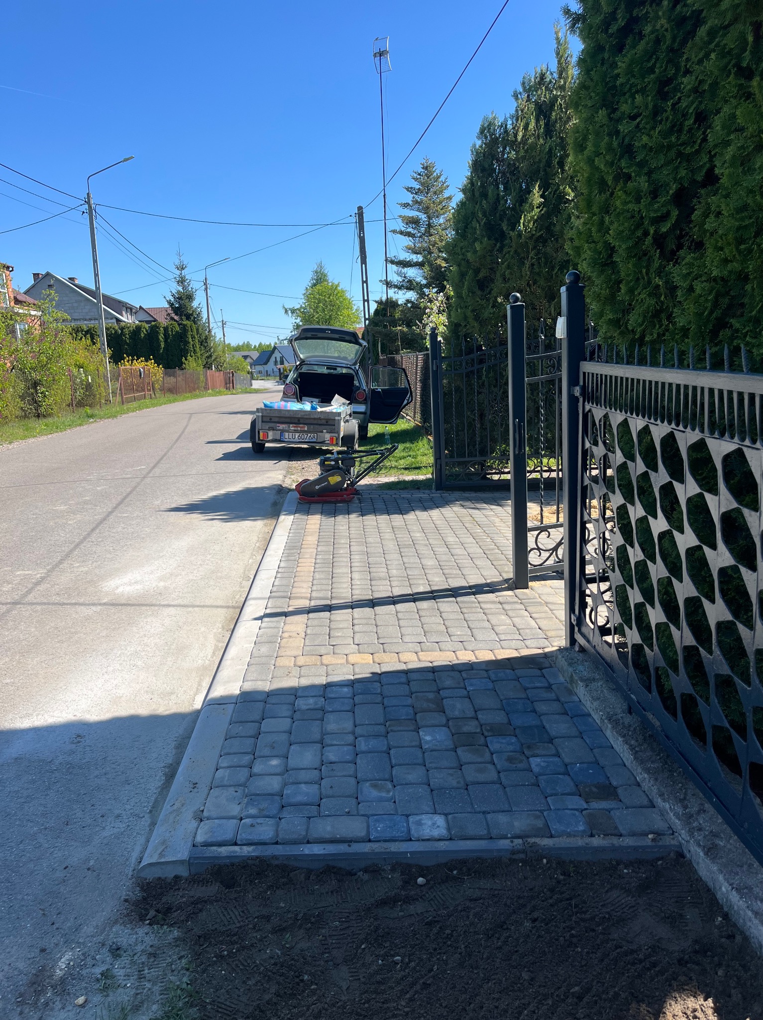 Ułożona kostka brukowa przed posesją z czarną bramą. W tle samochód z przyczepką i ubijarka. Widok na ulicę i domy w słoneczny dzień.