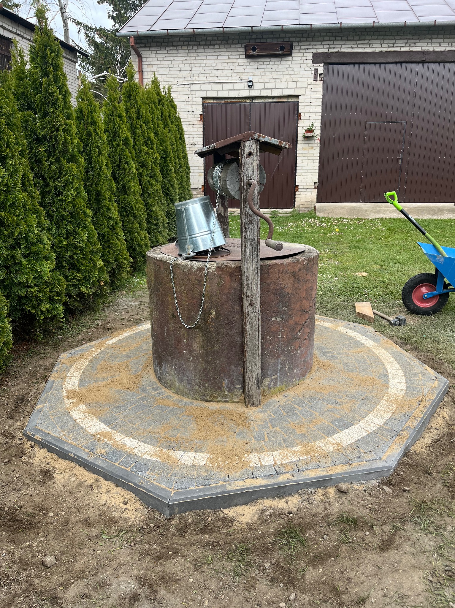 Studnia otoczona nową, szarą kostką brukową ułożoną w okrągły wzór, z elementami jasnego piasku i widocznym fragmentem wózka ogrodowego w tle.