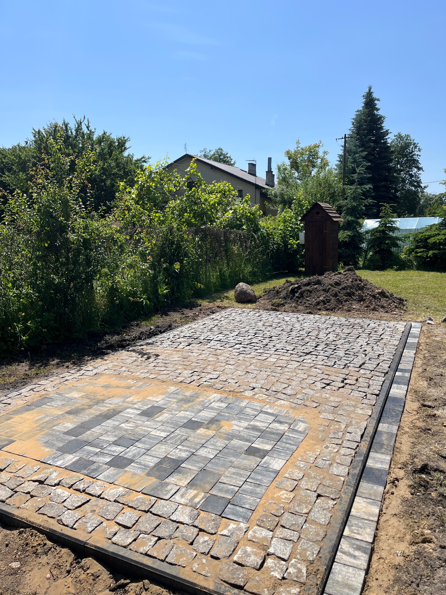 Fragment układanej kostki brukowej w ogrodzie, z widocznym obrzeżem i piaskiem, na tle zieleni i małej drewnianej budki. Słoneczny dzień.