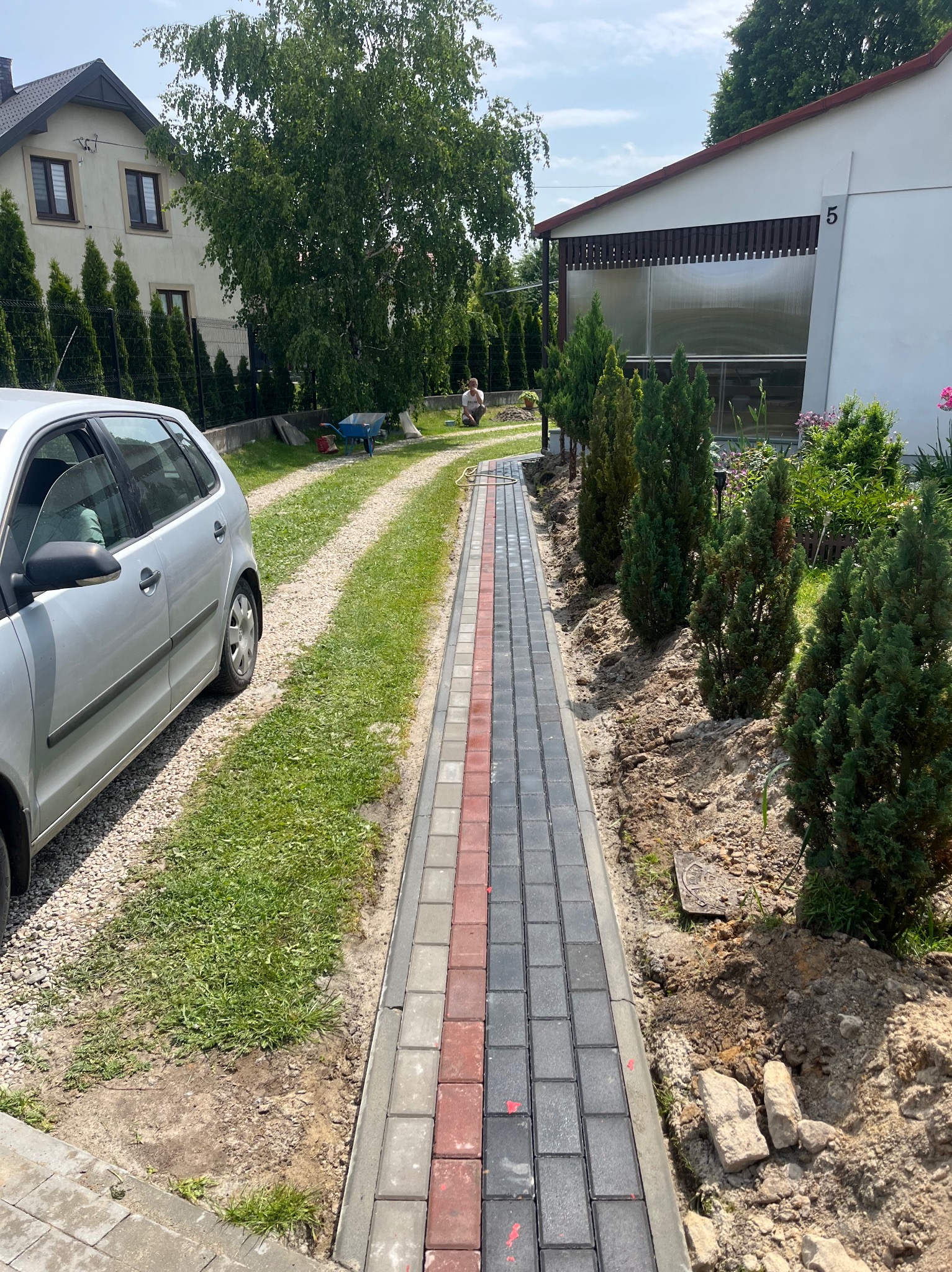 Ułożona ścieżka z kostki brukowej w odcieniach szarości i czerwieni, prowadząca przez trawnik w kierunku domu. W tle widoczny samochód i osoba pracująca w ogrodzie.