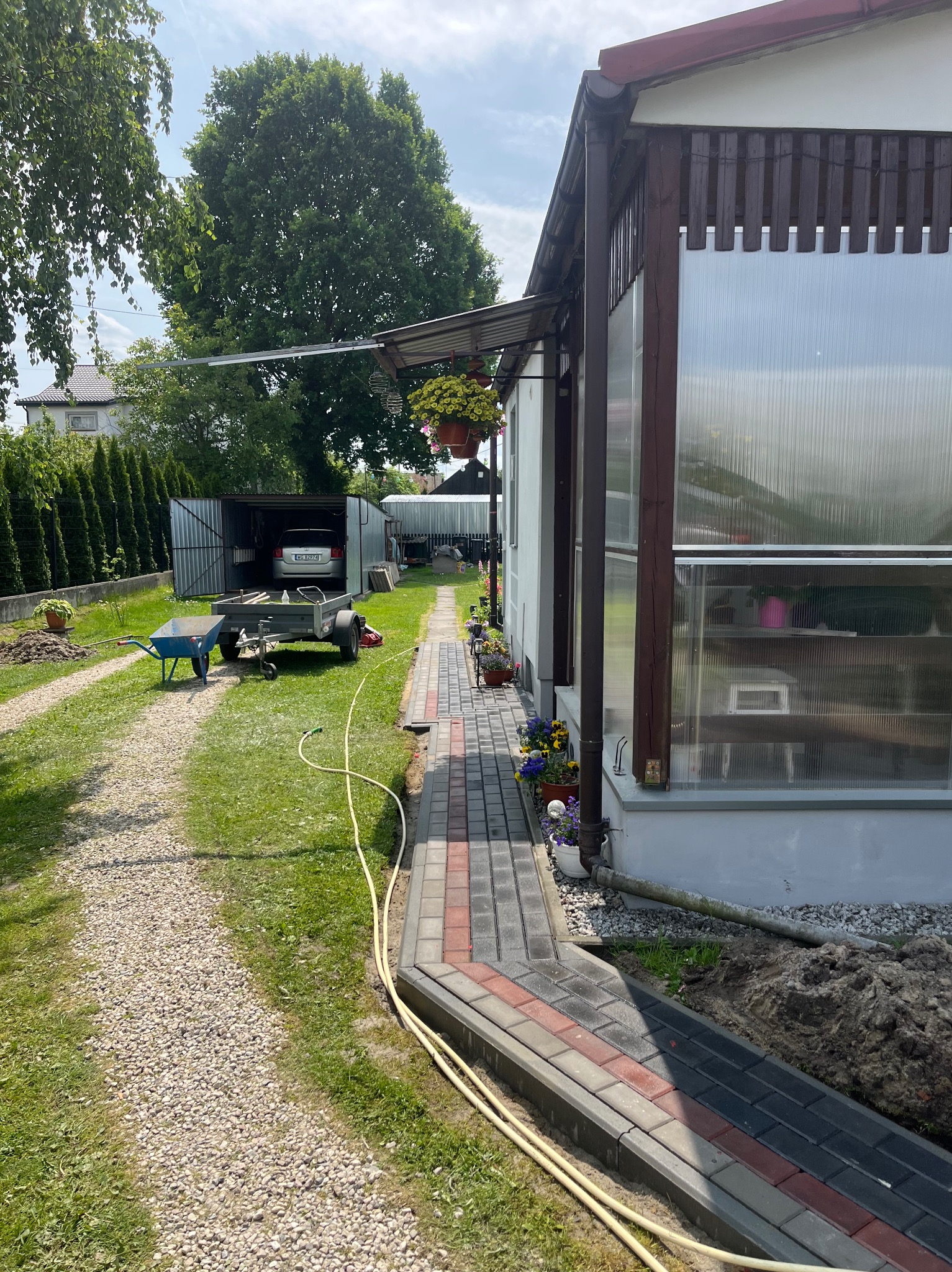 Ogród z ścieżką z kostki brukowej w odcieniach szarości i czerwieni, prowadzącą do szklarni. Widoczny wózek, przyczepka i samochód w garażu.