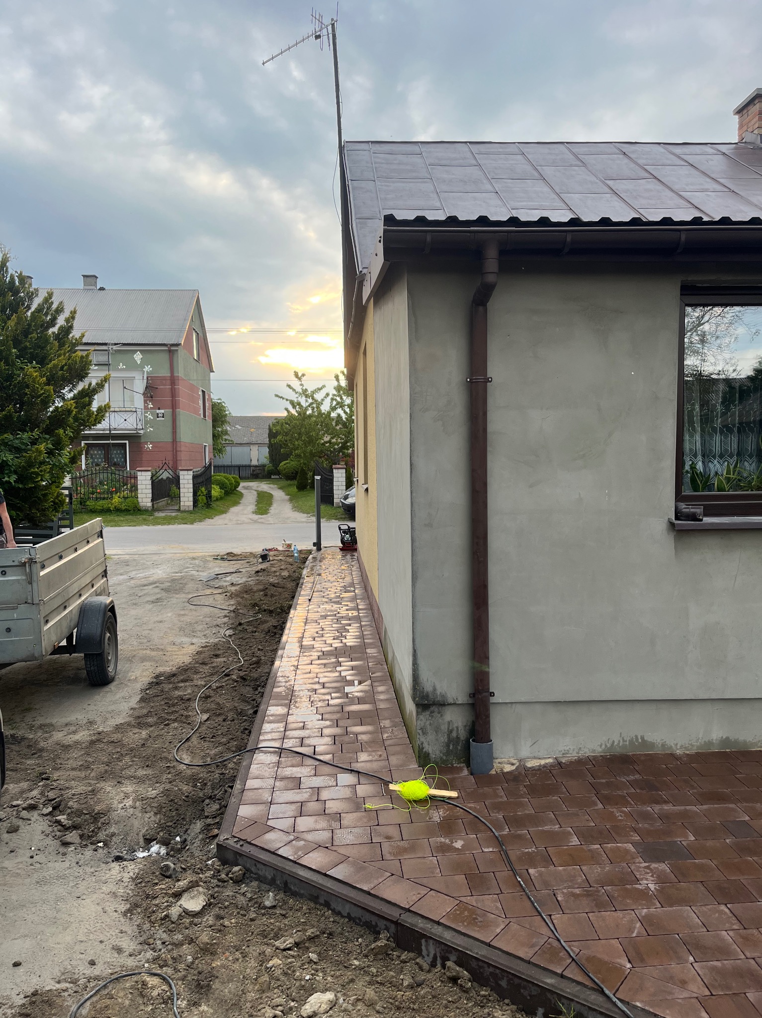 Brązowa kostka brukowa ułożona wzdłuż ściany domu, widoczne narzędzia i ziemia. W tle dom i zachodzące słońce.
