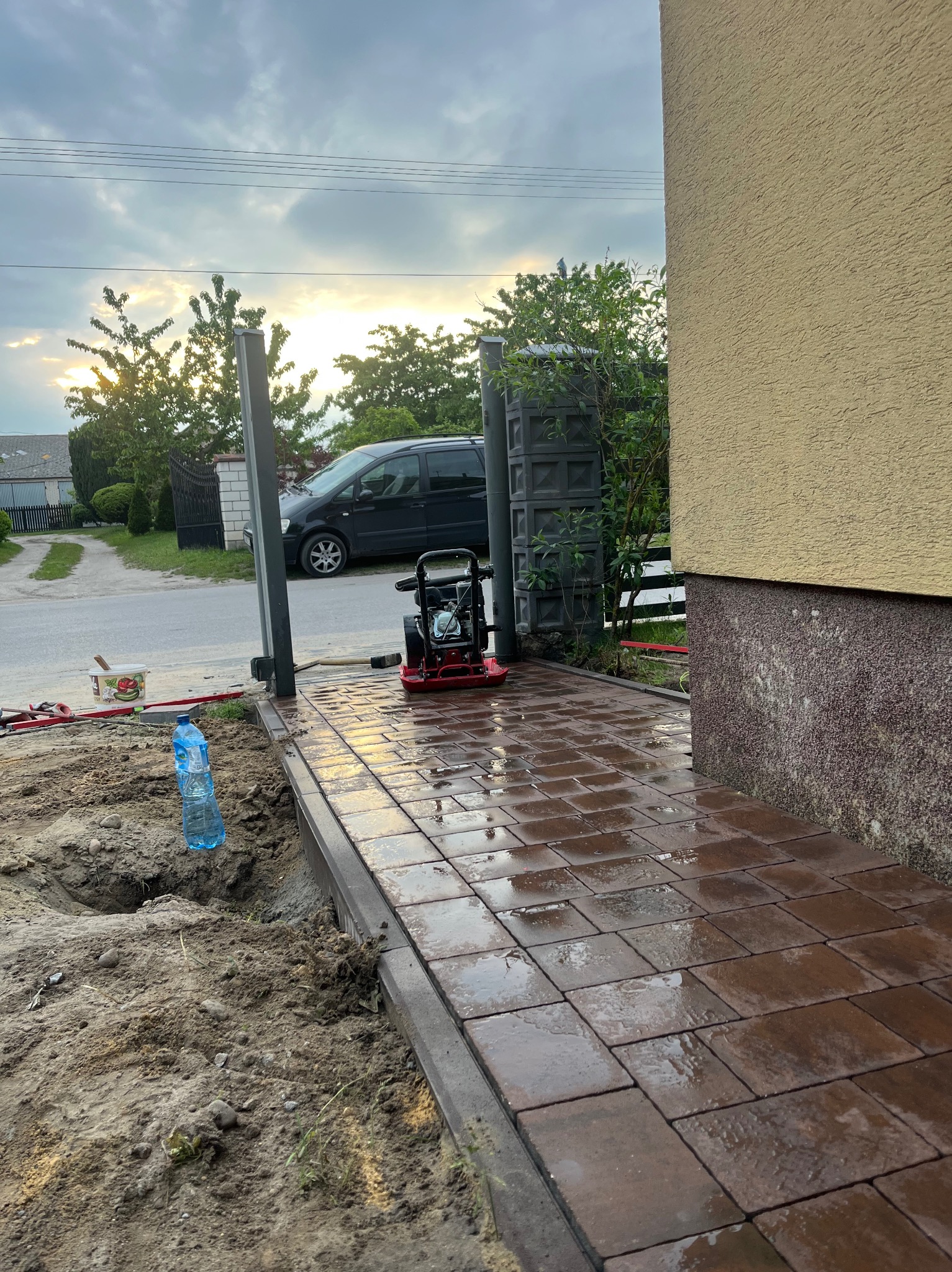 Świeżo ułożona, mokra kostka brukowa w odcieniach brązu, z widoczną zagęszczarką i wykopem obok. W tle brama, samochód i zachmurzone niebo.