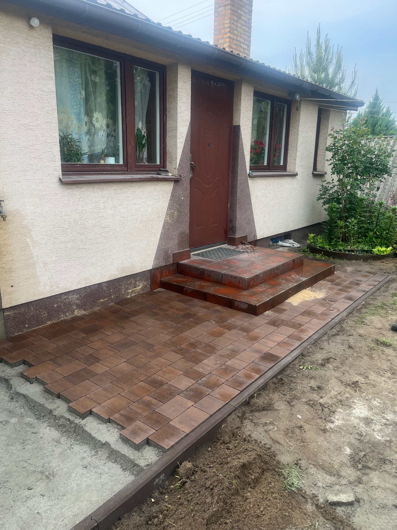 Ułożona, brązowa kostka brukowa prowadzi do wejścia domu z dwoma stopniami. Widoczne niedokończone krawędzie i świeżo ułożona nawierzchnia.