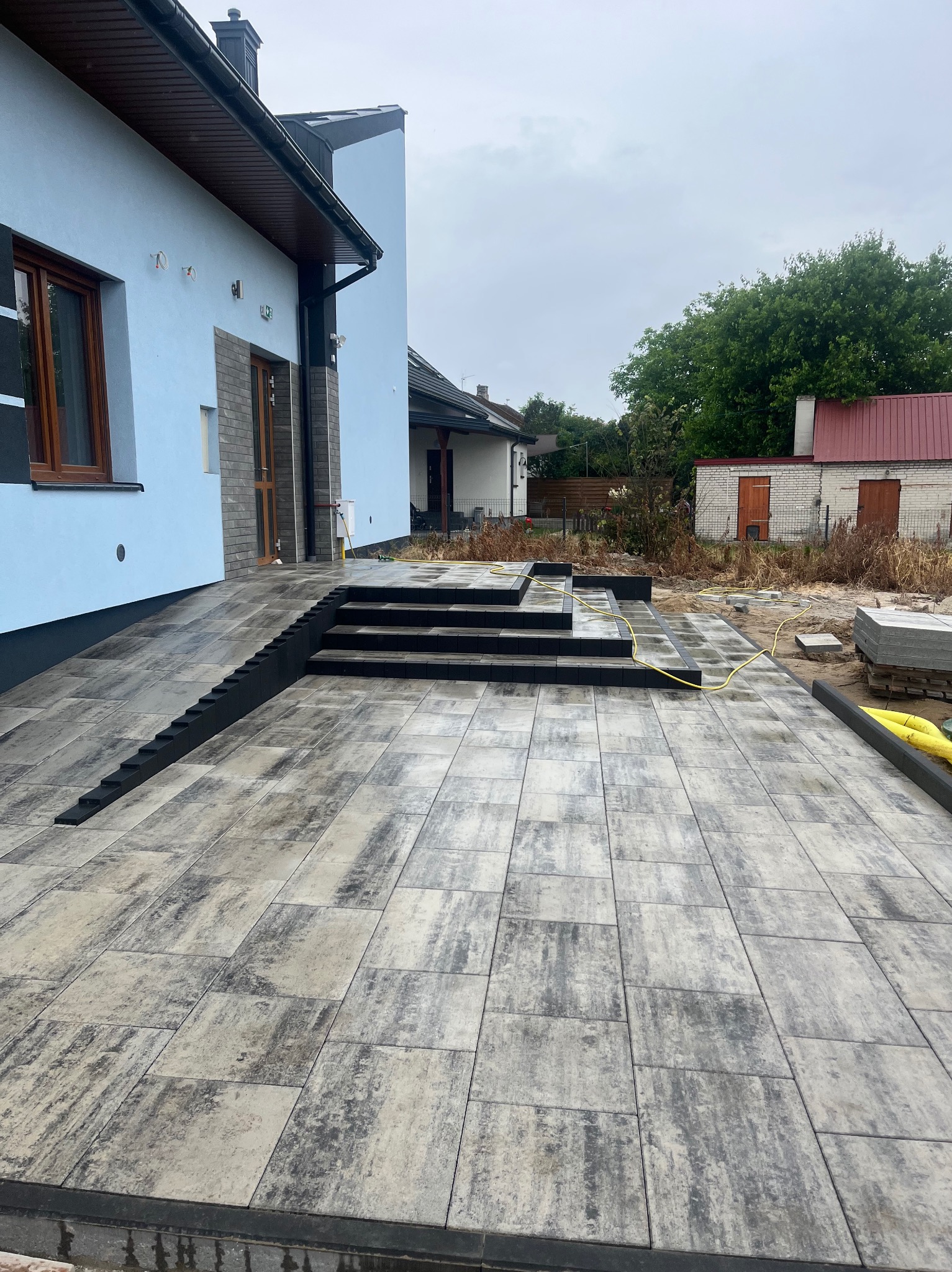 Ułożona kostka brukowa w odcieniach szarości prowadząca do wejścia domu z rampą i schodami. Widoczne palety z kostką i elementy wykończeniowe.