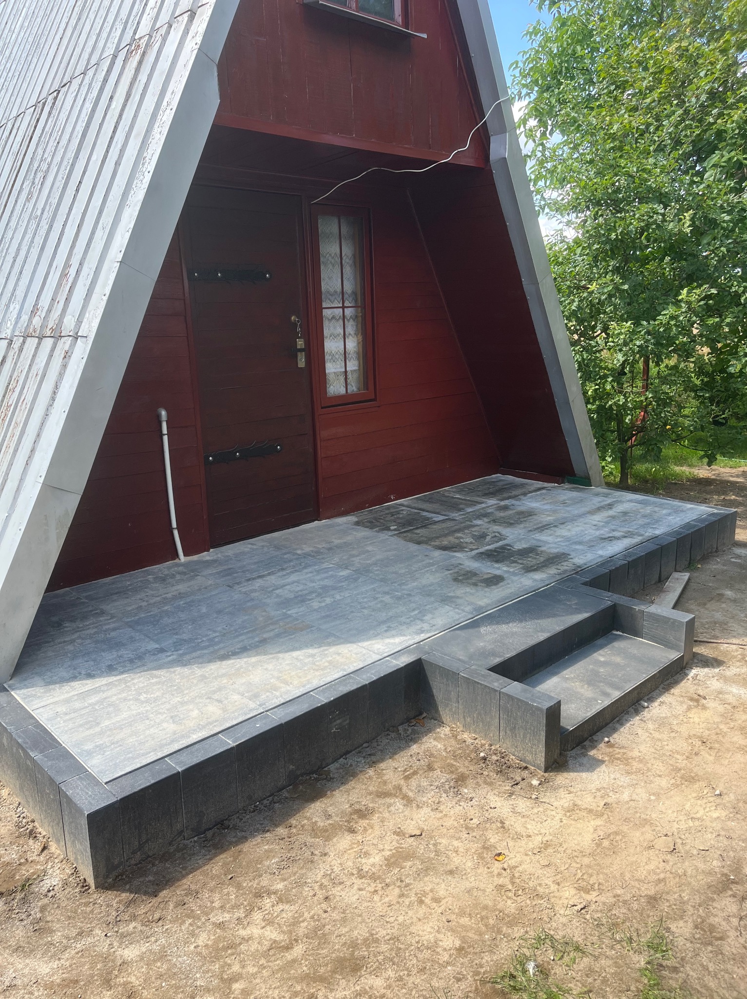 Nowoczesny taras z szarej kostki brukowej przed domkiem w stylu A-frame. Schody wejściowe wykończone tą samą kostką. Widok na brązowe drzwi wejściowe.