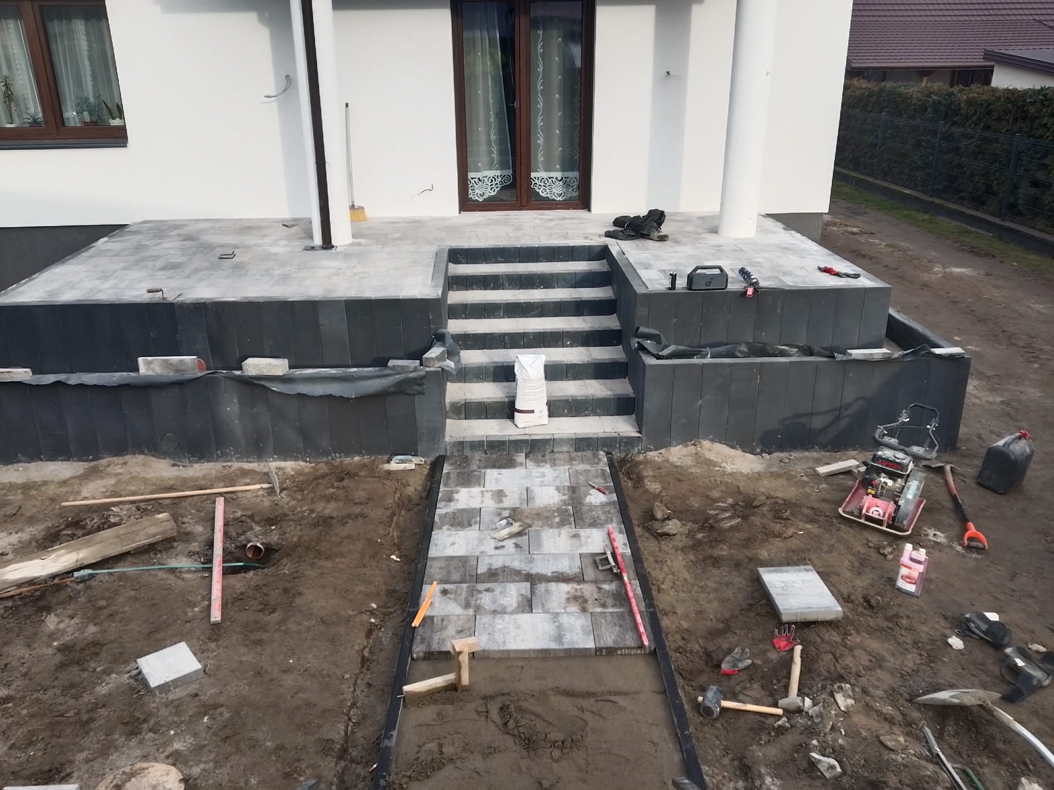 Etap budowy schodów i ścieżki z kostki brukowej przed domem. Widoczne narzędzia, materiały i częściowo ułożona kostka. Ujęcie z góry, w tle biały budynek.