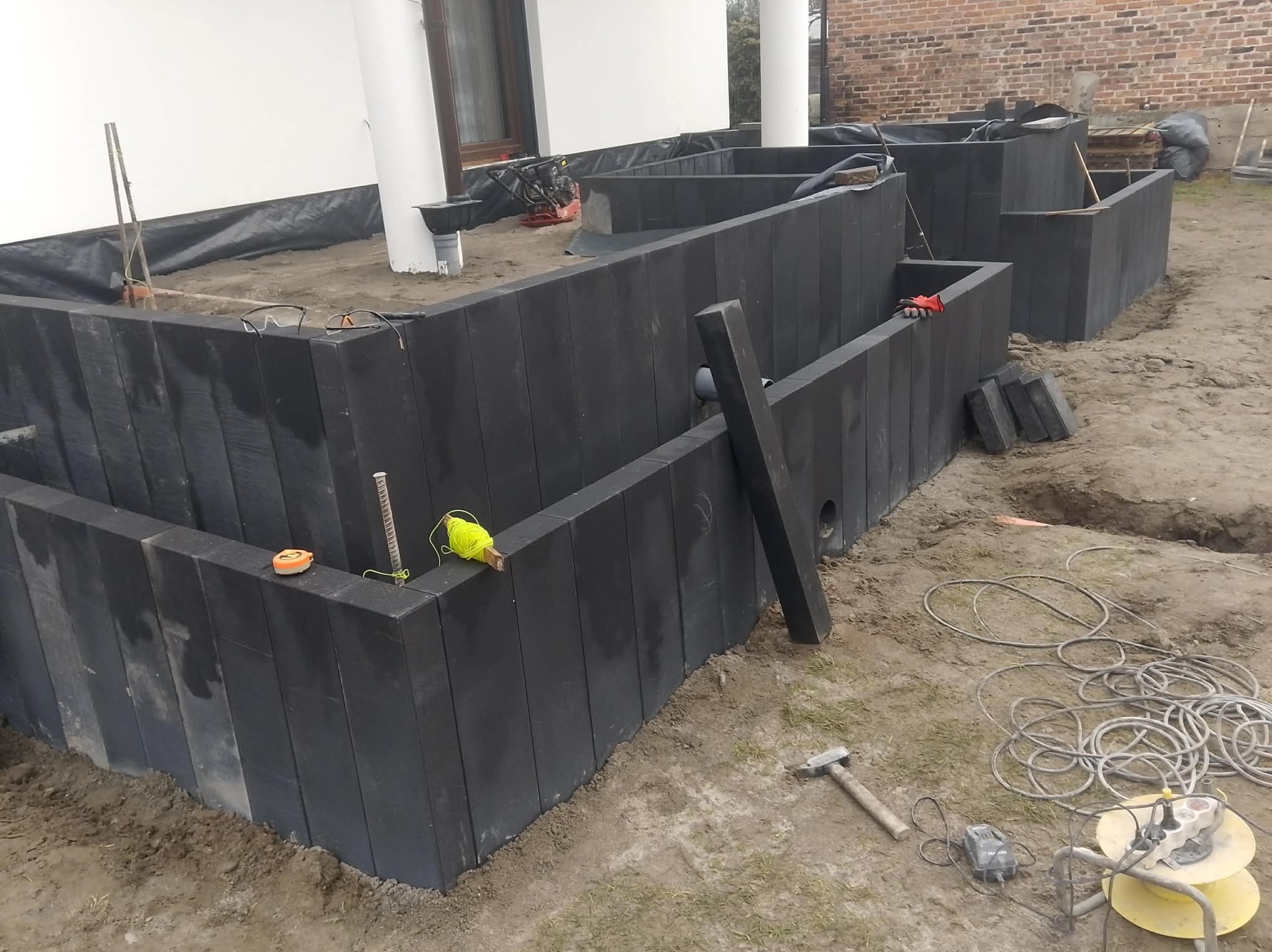 Montaż obrzeży rabat z ciemnych, pionowo ułożonych elementów. Widoczne narzędzia: młotek, poziomica, sznurek traserski. Częściowo wyłożona folia. Etap prac ziemnych.