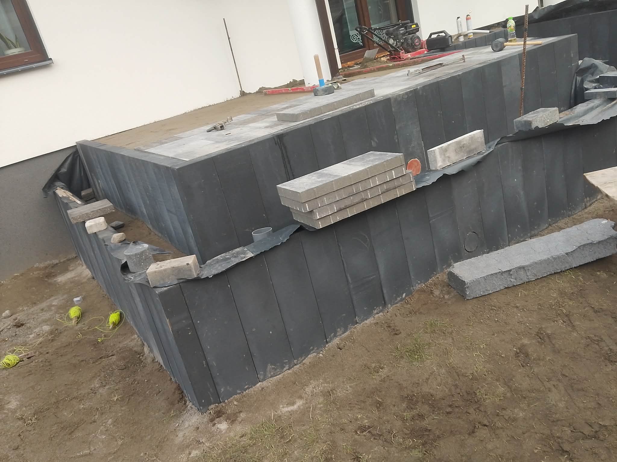 Budowa tarasu z kostki brukowej: czarne bloczki fundamentowe, szara kostka, zagęszczarka i narzędzia na placu budowy. Widok z dołu, częściowo ukończony etap.