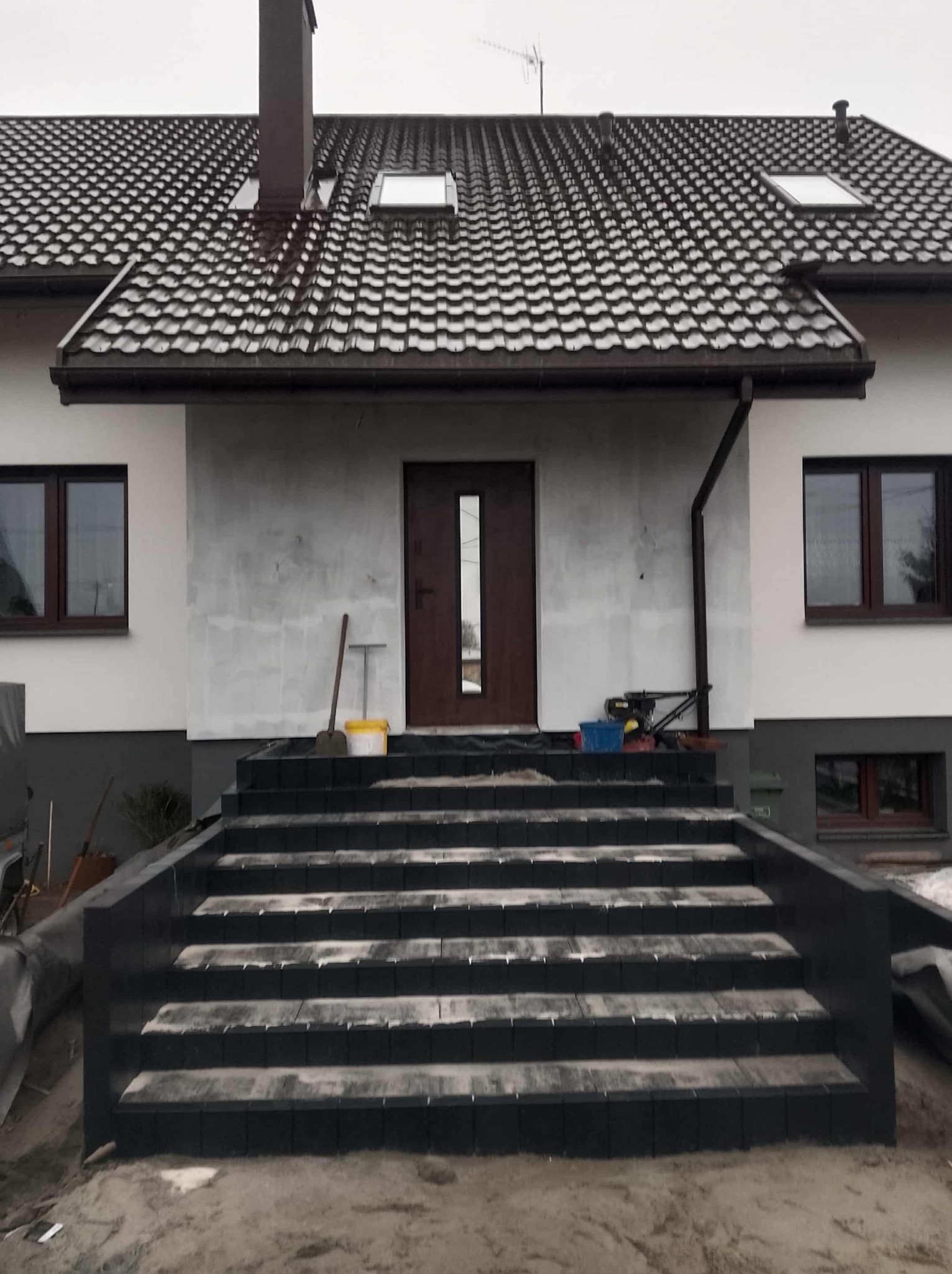 Dom z nowymi, ciemnymi schodami wejściowymi z kostki brukowej. Ściana frontowa w trakcie wykończenia, widoczne narzędzia i materiały budowlane.
