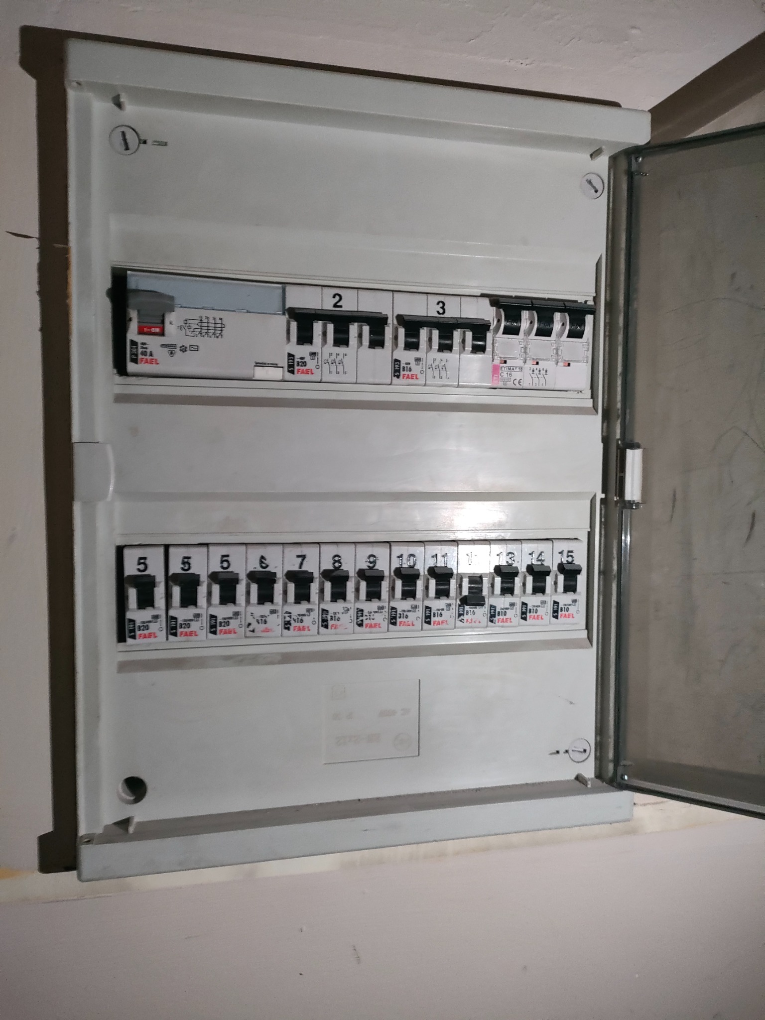 Otwarta skrzynka rozdzielcza z bezpiecznikami FAEL, widoczne numery bezpieczników od 1 do 15, na tle białej ściany.