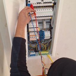 SILESIA INSTAL Tomasz Kulawik - Elektryk mierzy napięcie w skrzynce rozdzielczej z otwartymi drzwiczkami, używając multimetru. Widoczne kable, bezpieczniki i inne elementy instalacji elektrycznej.