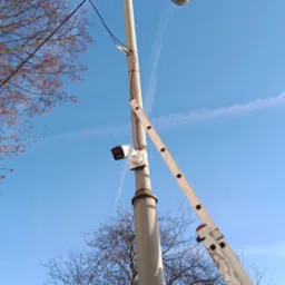 Montaż kamery monitoringu na słupie oświetleniowym. Przystawiona drabina, błękitne niebo w tle. Widok z dołu na zainstalowaną kamerę.