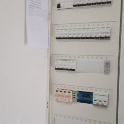 SILESIA INSTAL Tomasz Kulawik - Otwarta, biała skrzynka rozdzielcza z bezpiecznikami i listwą zaciskową, z numeracją 1-60, umieszczona wewnątrz budynku. Na drzwiach skrzynki widoczna kartka z listą.