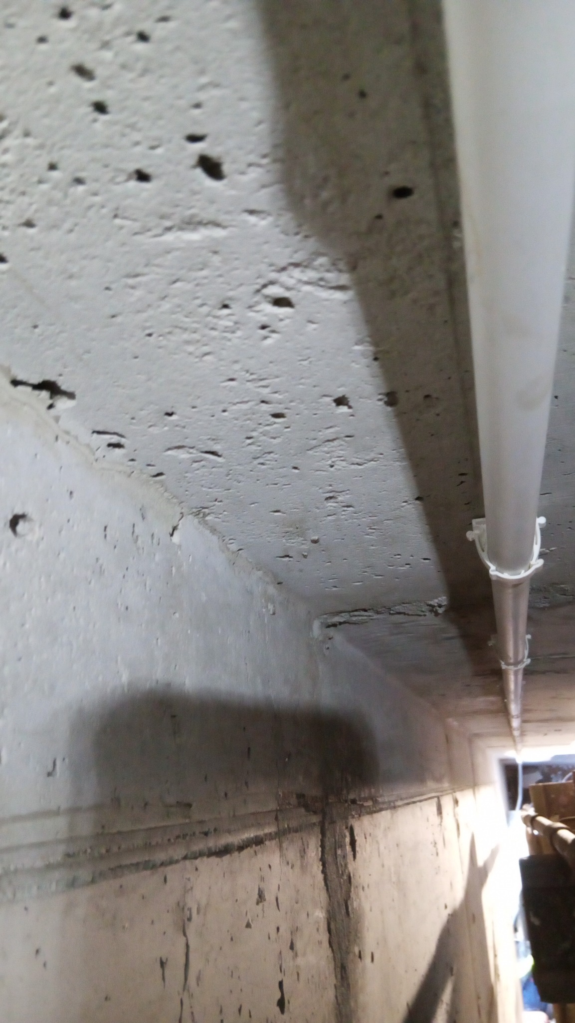 Biała rura przymocowana do surowego, betonowego sufitu w pomieszczeniu. Widoczne ślady wilgoci i niedoskonałości betonu. Niski kąt ujęcia.