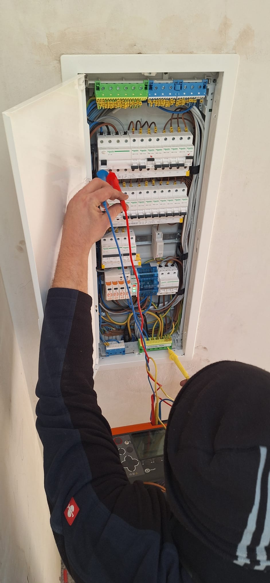 Elektryk mierzy napięcie w skrzynce rozdzielczej z otwartymi drzwiczkami, używając multimetru. Widoczne kable, bezpieczniki i inne elementy instalacji elektrycznej.