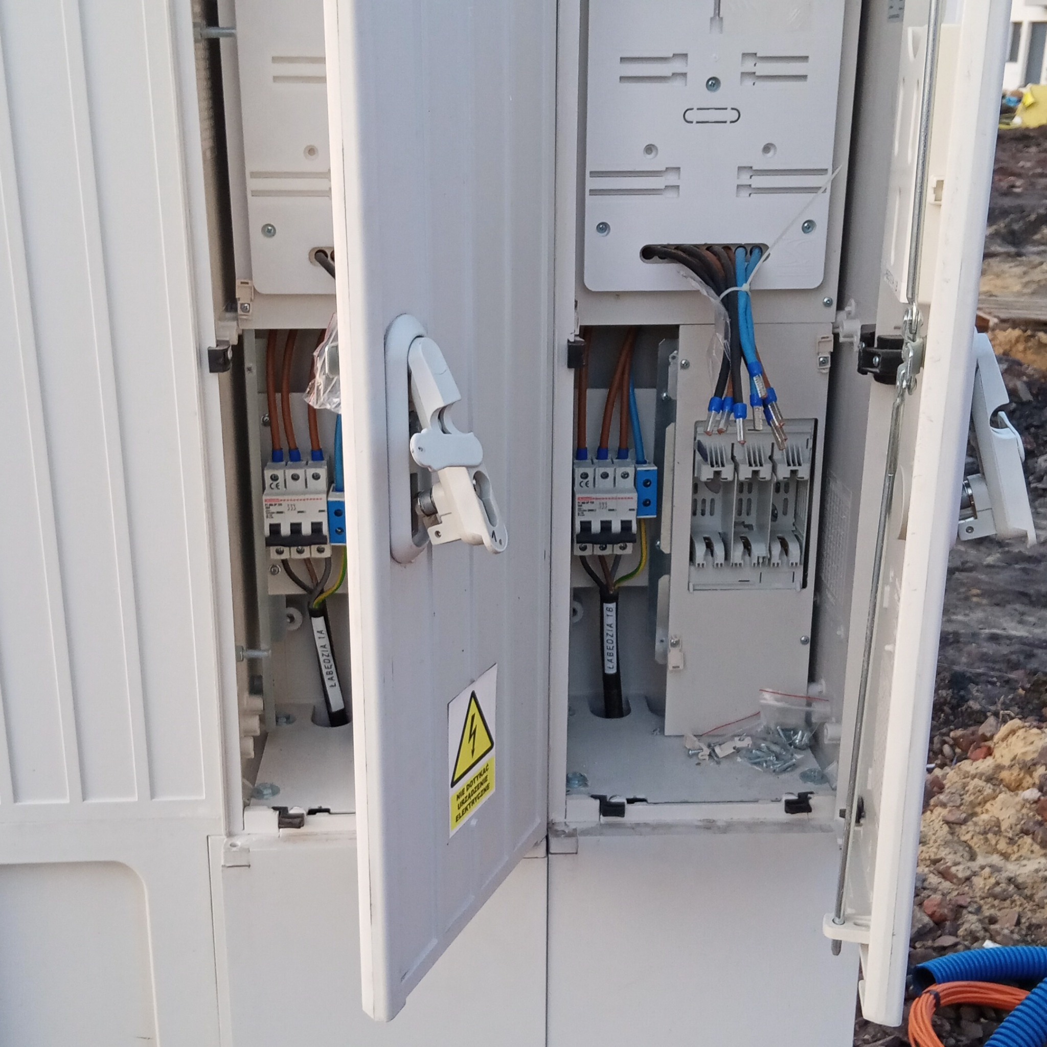 Otwarta skrzynka elektryczna z widocznymi bezpiecznikami, przewodami i elementami montażowymi, z żółtym znakiem ostrzegawczym na drzwiach.