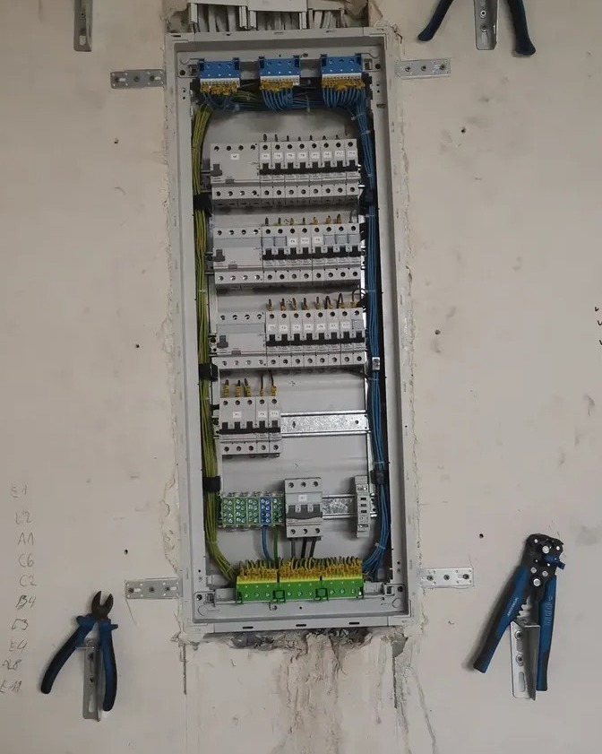 Rozdzielnica elektryczna w trakcie montażu w surowym wnętrzu. Widoczne przewody, bezpieczniki i narzędzia monterskie. Ściana z widocznymi śladami po instalacji.
