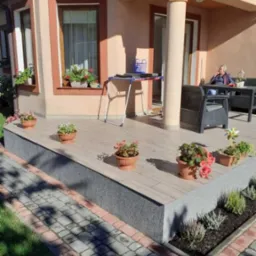 Zadaszony taras z drewnopodobnych paneli, ozdobiony doniczkami z kwiatami, widok z poziomu chodnika z kostki brukowej