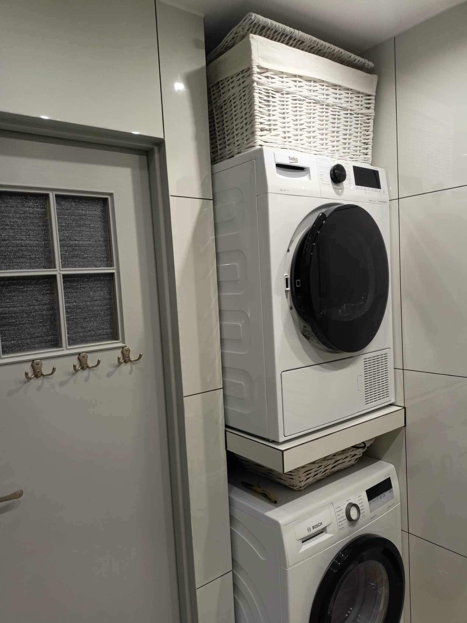 Pralka i suszarka ustawione jedna na drugiej w łazience z szarymi płytkami, biała wiklinowa skrzynka na górze, drzwi z wieszakami po lewej stronie.