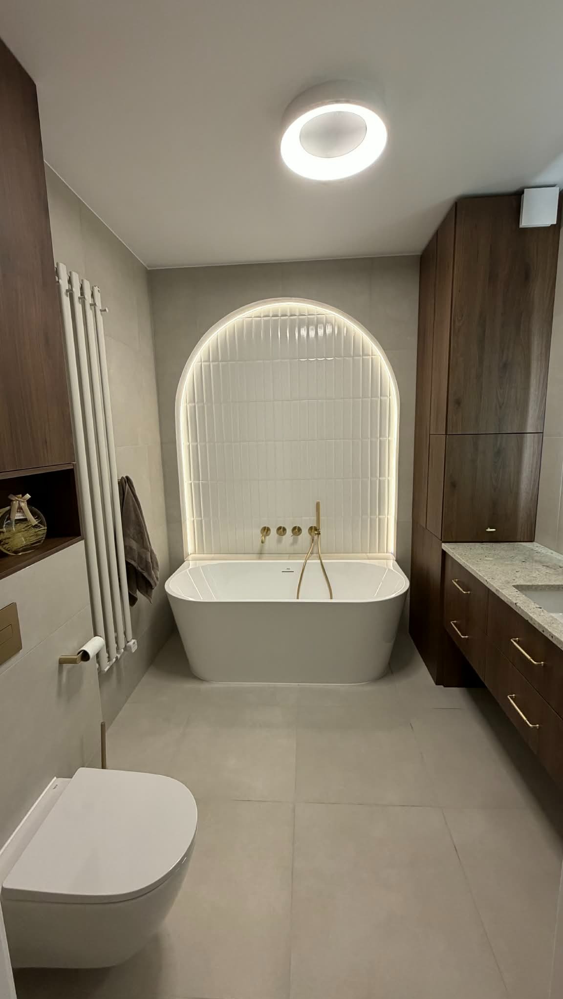 Elegancka łazienka z wolnostojącą wanną na tle białych, podświetlanych płytek w kształcie łuku. Umywalka z szafką z ciemnego drewna i złotymi uchwytami. Minimalistyczny grzejnik.