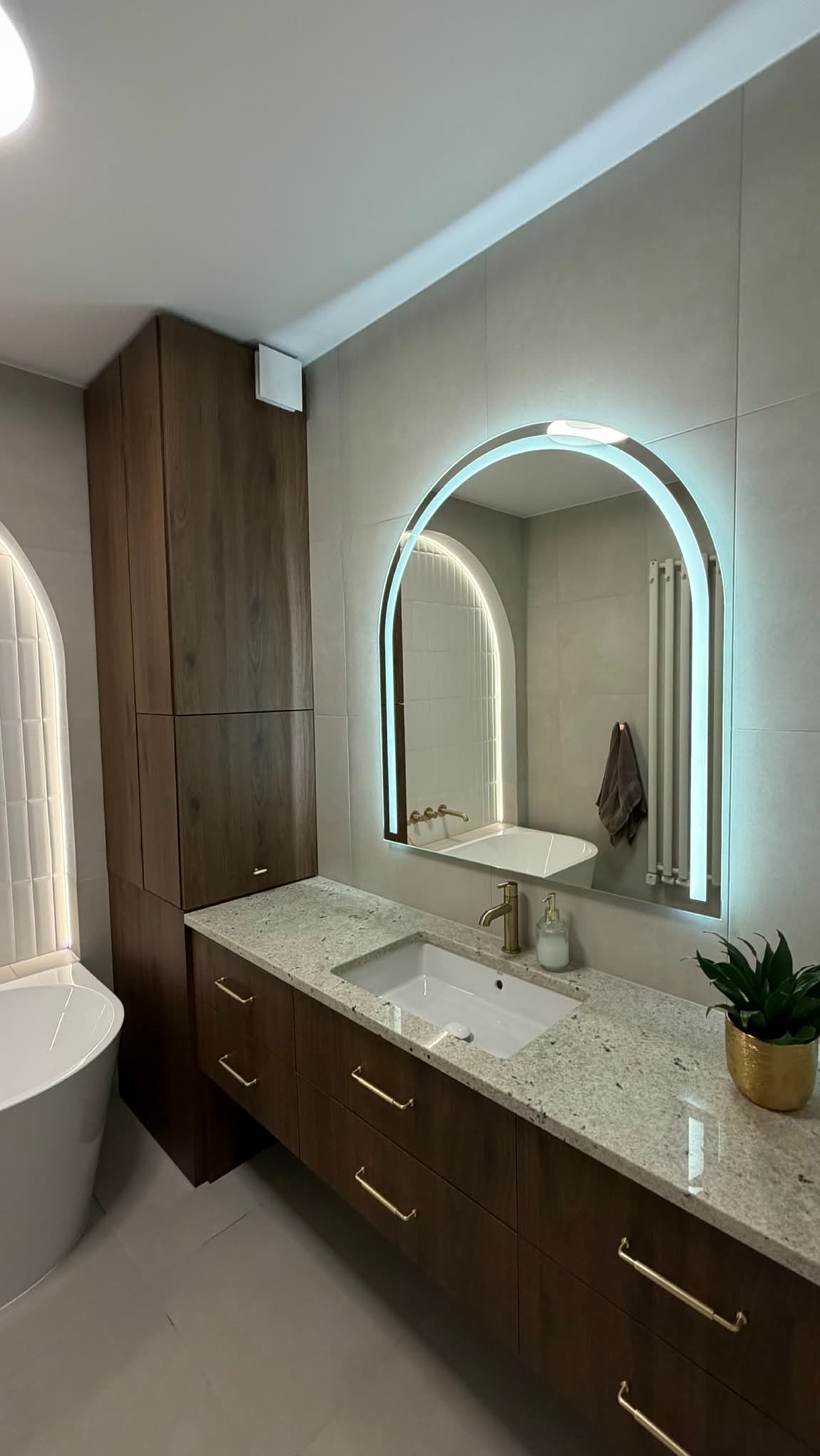 Nowoczesna łazienka z podświetlanym lustrem w kształcie łuku, drewnianą szafką z granitowym blatem i złotymi uchwytami. Widoczna wanna i roślina w złotej doniczce.