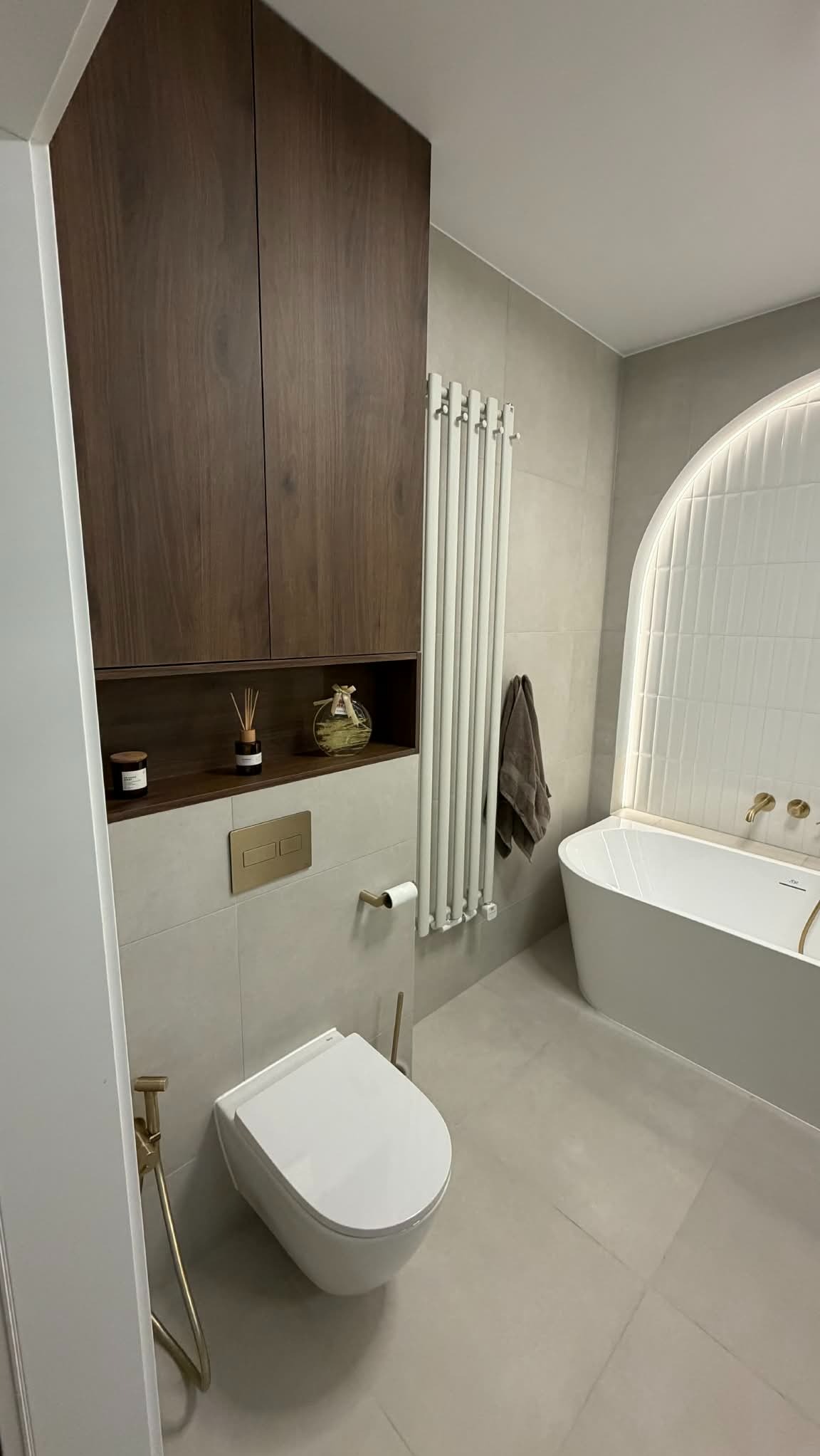 Nowoczesna łazienka z wiszącą toaletą, brązową szafką i grzejnikiem drabinkowym. Uwagę zwraca wanna z podświetlaną, łukową wnęką z białych płytek.