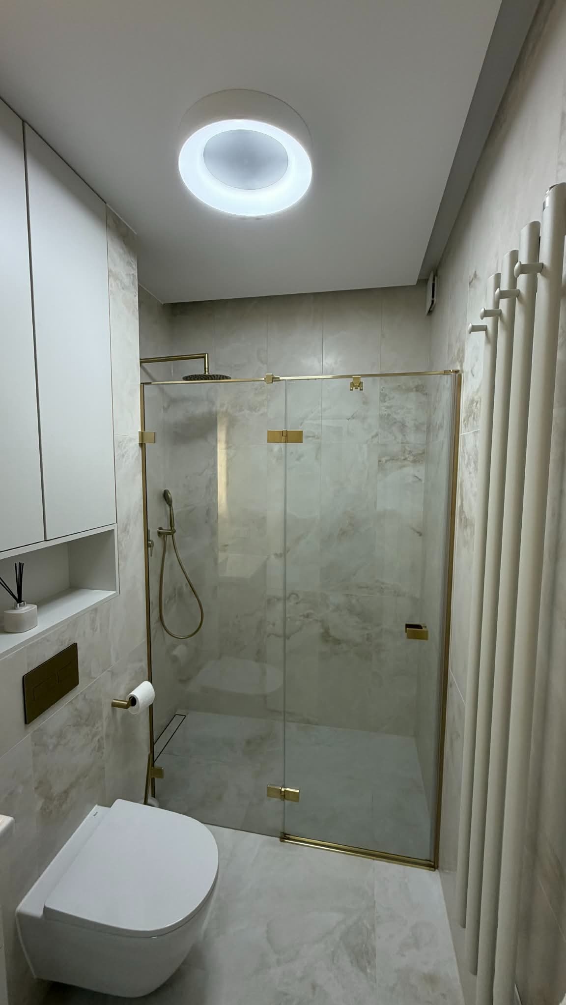 Wykończona łazienka z kabiną prysznicową ze złotymi detalami, wiszącą toaletą i grzejnikiem drabinkowym. Płytki imitujące marmur na ścianach i podłodze.