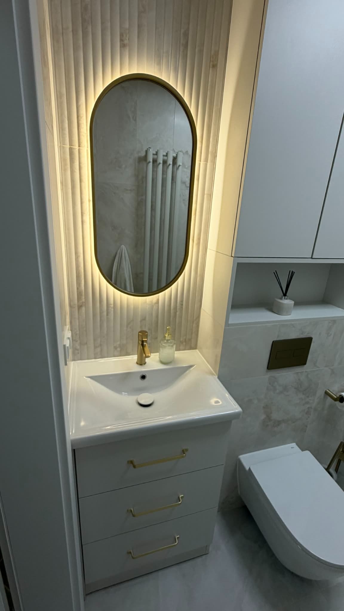 Nowoczesna łazienka z owalnym lustrem podświetlanym LED, białą umywalką z szafką i złotymi akcentami. Widoczna toaleta i zabudowa meblowa w jasnych barwach.