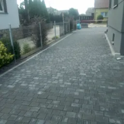 Ułożona szara kostka brukowa na podjeździe, widoczne ogrodzenie z siatki, budynek w tle, częściowo ułożone obrzeża przy budynku.