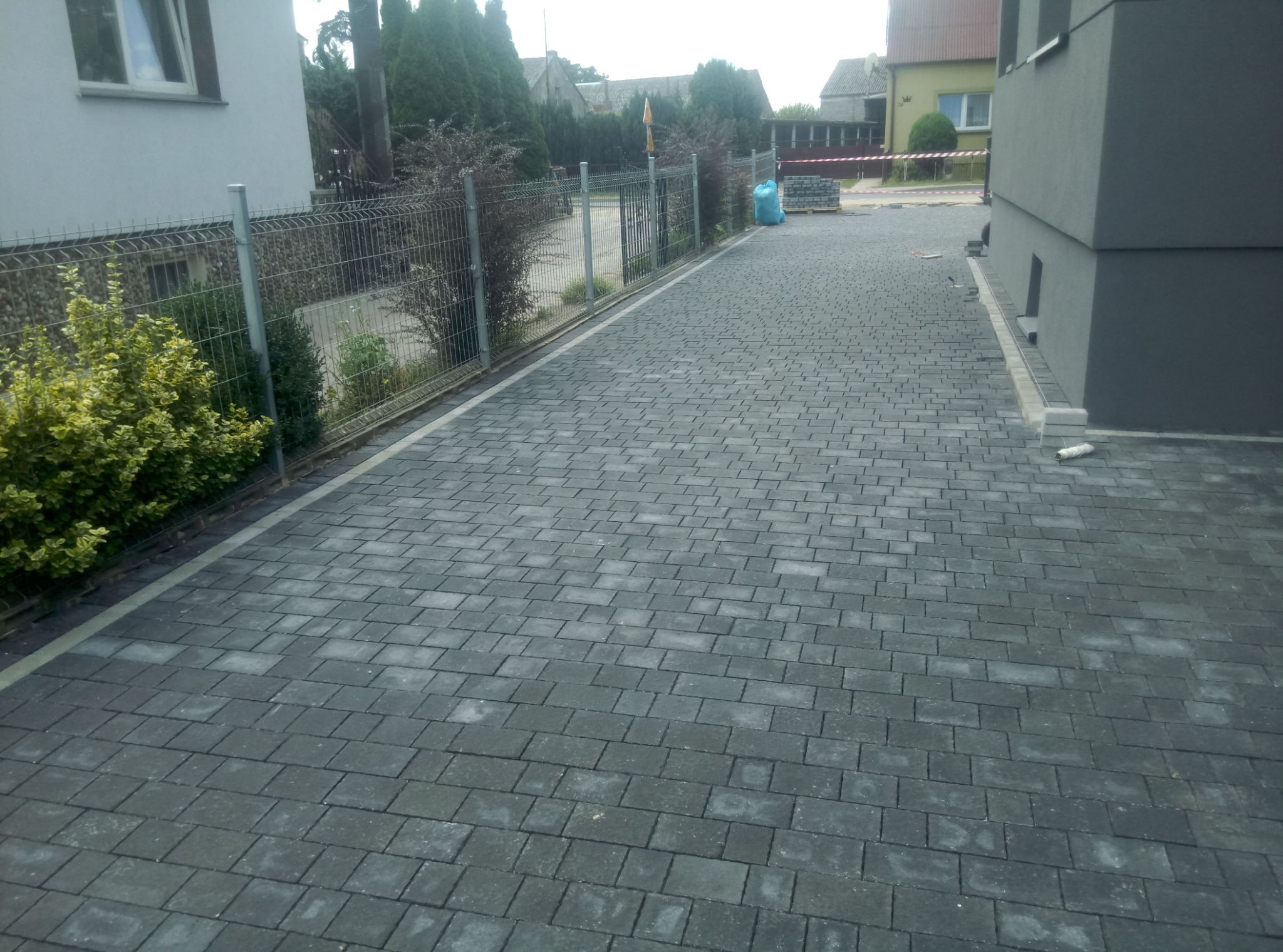 Ułożona szara kostka brukowa na podjeździe, widoczne ogrodzenie z siatki, budynek w tle, częściowo ułożone obrzeża przy budynku.