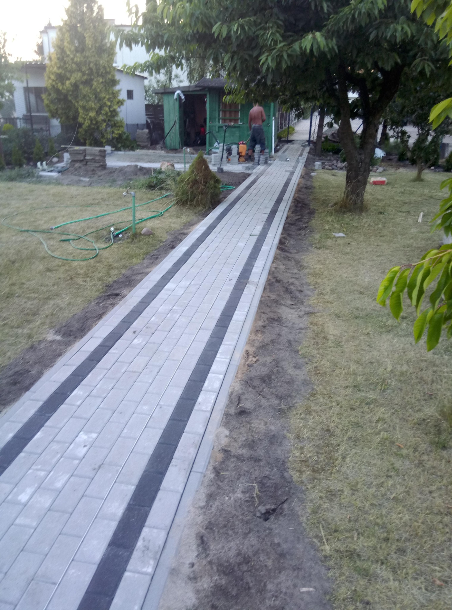 Ułożony chodnik z jasnoszarej kostki brukowej z ciemnymi pasami, widoczny mężczyzna przy pracy i narzędzia w tle, ogród w trakcie prac wykończeniowych.