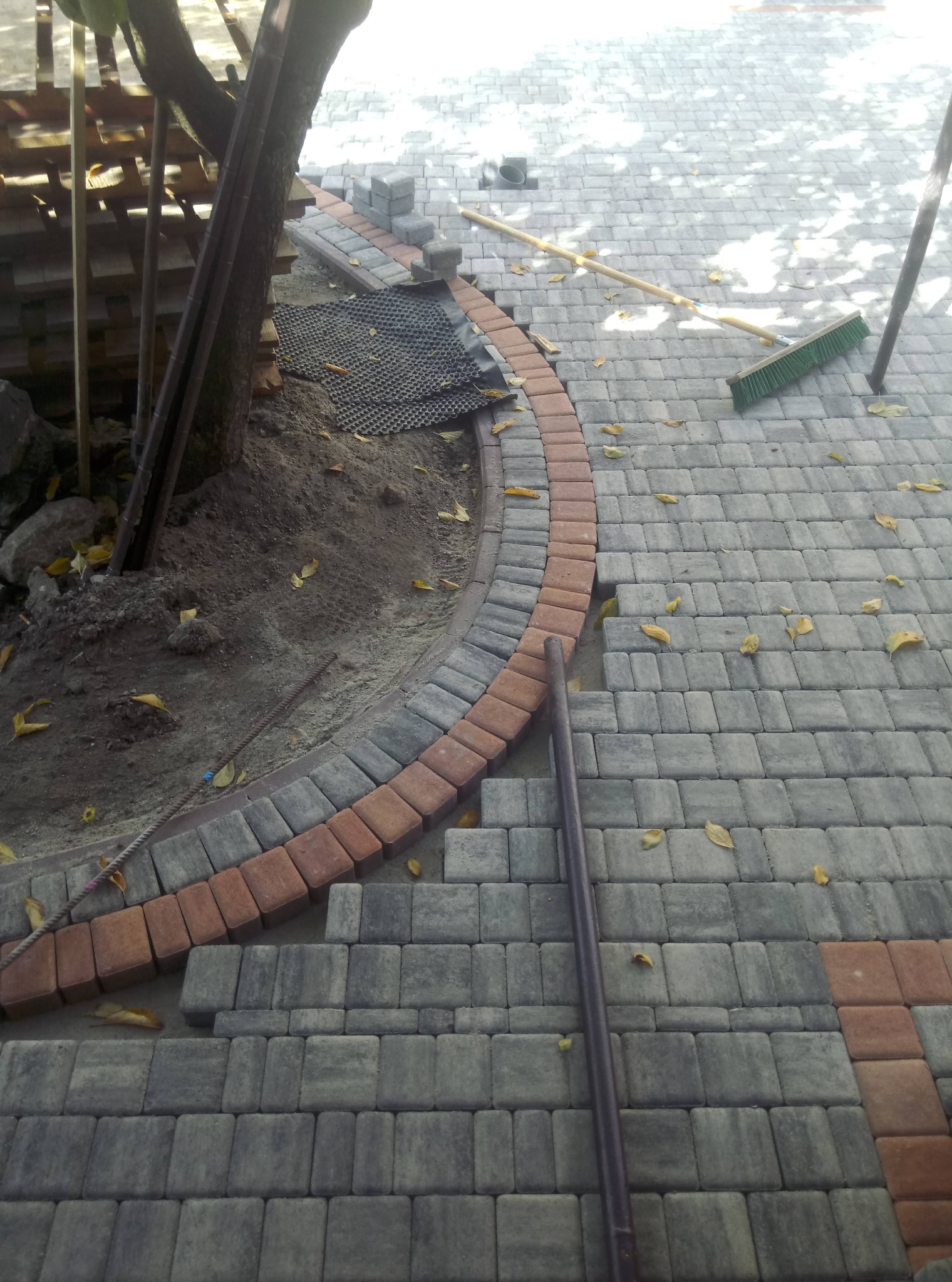 Fragment układanej nawierzchni z kostki brukowej w odcieniach szarości i ceglastej czerwieni, z widoczną obwódką wokół drzewa i przygotowanym podłożem, częściowo zasypanym liśćmi. Obok leży miotła...