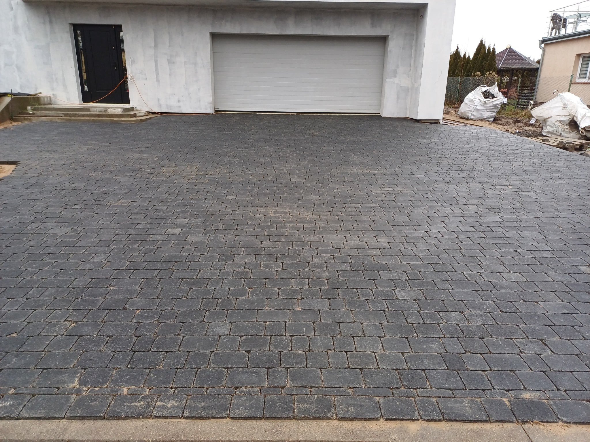 Ułożona ciemnoszara kostka brukowa na podjeździe przed nowym domem z białą elewacją i garażem, widoczne worki z odpadami budowlanymi z boku.