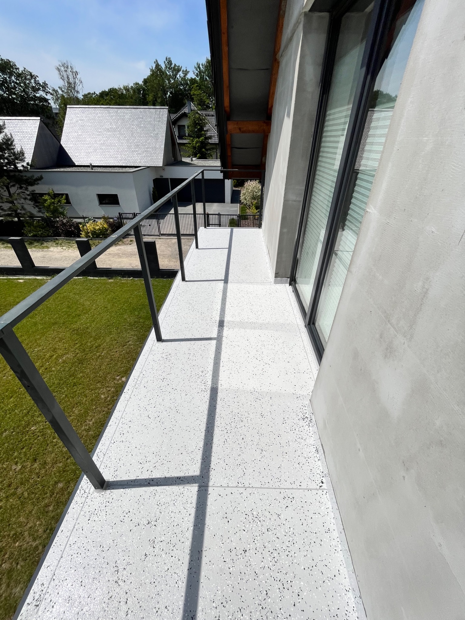 Biała powierzchnia balkonu z szarymi i czarnymi drobinkami, otoczona szarą metalową balustradą i betonową ścianą budynku, z widokiem na domy z szarymi dachami i zielony trawnik w tle.