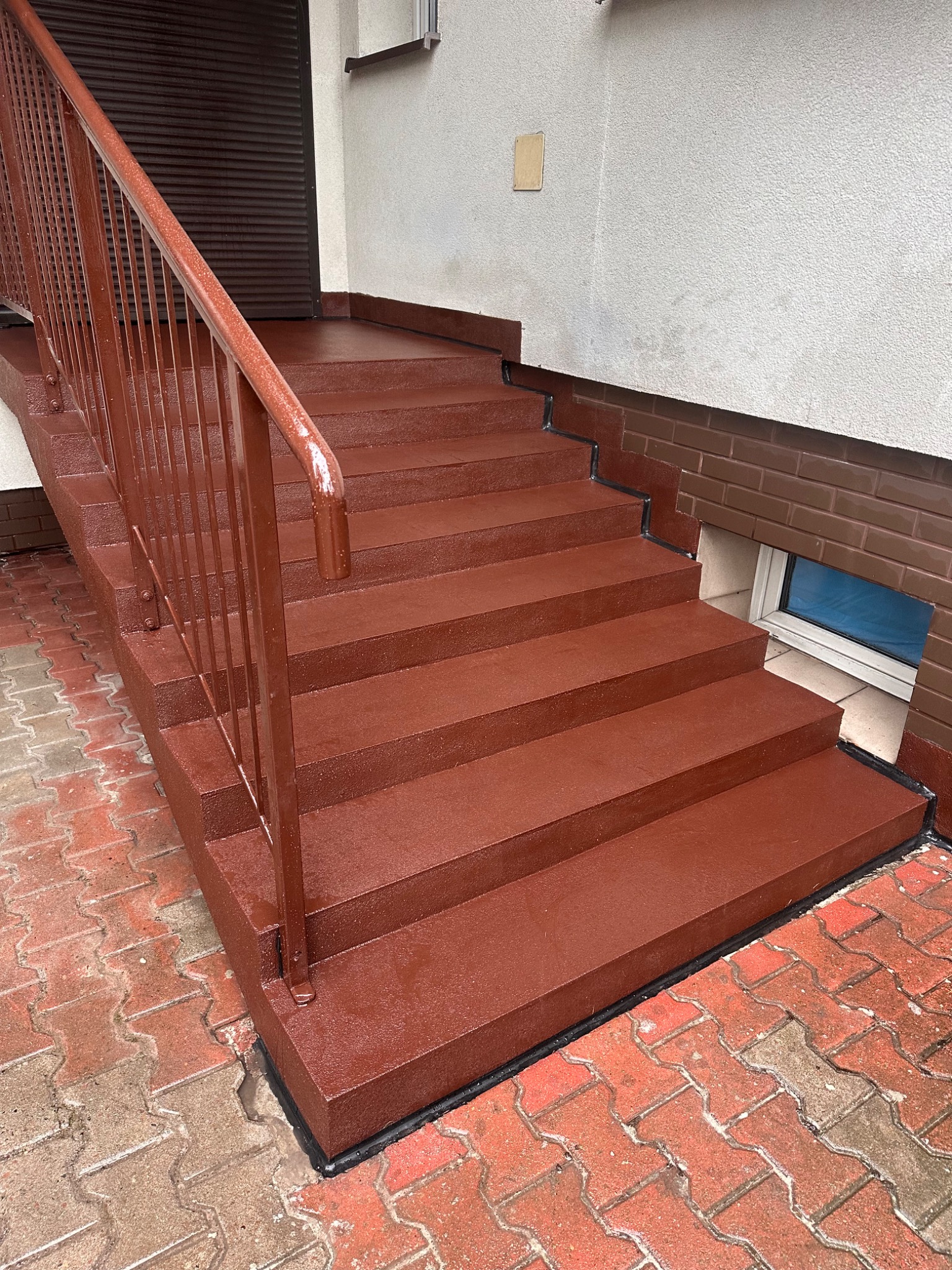 Schody zewnętrzne pokryte jednolitą, brązową posadzką żywiczną. Widoczne metalowe balustrady w kolorze posadzki oraz fragment chodnika z kostki brukowej. Na ścianie w tle widoczny fragment okna...